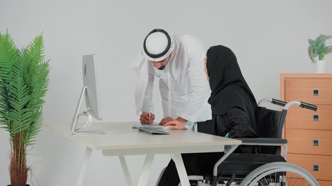 Dialogue and discussion between colleagues, comfortable work environment in Saudi companies, a veiled Arab Gulf woman sitting in a wheelchair exchanging experiences and information to achieve goals, an Arab Gulf Emirati man wearing a kandura and ghutra writing notes with his colleague, the use of computers and technology in the business field