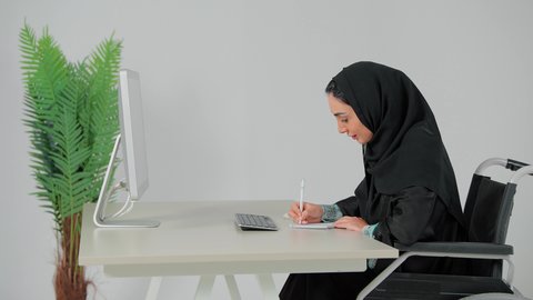 انجاز المهام اليومية، كتابة تقارير وأبحاث علمية ، استخدام الورق الأبيض في تسجيل الملاحظات والمعلومات المهمة، مفهوم كتابه وتدوين الملاحظات، تصوير من الجانب لامراة عربية خليجية سعودية تجلس على كرسي متحرك امام الحاسب الالى تمسك بيدها قلم تقوم بالكتابة على نوت