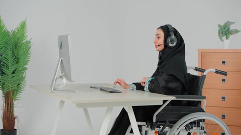 الرد على الاستعلامات والاستفسارات، النقر على لوحة المفاتيح ، مكالمات فيديو خلال اجتماع العمل, إمرأة عربية خليجية سعودية محجبة ترتدي عباءة سوداء تجلس على كرسي متحرك تضع سماعات الهيدفون تعمل في مجال خدمة العملاء، استخدام التقنيات الحديثة والمتطورة