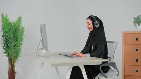 النقر على لوحة المفاتيح ، مكالمات فيديو خلال اجتماع العمل, إمرأة عربية خليجية سعودية محجبة ترتدي عباءة سوداء تضع سماعات الهيدفون تعمل في مجال خدمة العملاء, الرد على الاستعلامات والاستفسارات، استخدام التقنيات الحديثة والمتطورة
