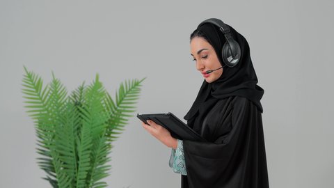 الاستماع عبر الهيدفون، إنجاز المهام من المنزل، دمج التقنية والتكنولوجيا في الروتين اليومي،  تصوير من الجانب لامراة عربيه خليجيه سعودية ترتدي العباءة و الحجاب و سماعات الاذن تقوم باستخدام جهاز التابلت ، تصفح مواقع التواصل الاجتماعي