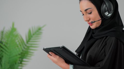 إنجاز المهام من المنزل، الاستماع عبر الهيدفون، دمج التقنية والتكنولوجيا في الروتين اليومي، تصوير مقرب من الجانب لامراة عربيه خليجيه سعودية ترتدي العباءة و الحجاب و سماعات الاذن تقوم باستخدام جهاز التابلت ، تصفح مواقع التواصل الاجتماعي