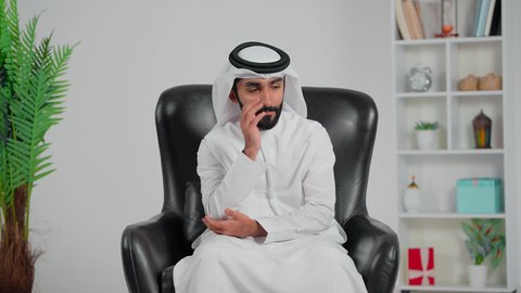 Finding a solution to a problem, expressions of distraction and contemplation, meditation and planning for the future, an Arab Gulf Emirati man wearing a kandura and ghutrah sitting on a chair with his hand on his chin in gestures of thinking and focus.