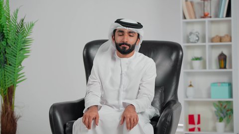 إيماءات الراحة والإسترخاء، ،رجل عربي خليجي اماراتي يرتدي الكندورة و الغتره يجلس على كرسي يضع يديه اسفل رأسه يغمض عينيه بتعابير النعاس، الشعور بالسعادة والسرور