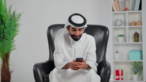 قضاء اوقات الفراغ الممتعة، مفهوم الاندماج بمشاهدة الفريق المفضل ، الحماس والتفاعل مع المنتخب ،المباريات والألعاب الرياضية، الحماس بالفوز بالكأس، رجل عربي خليجي اماراتي يرتدي الكندورة و الغتره يجلس على كرسي يمسك بيده الهاتف المحمول و يقوم بمشاهدة ومتابعة كرة القدم بتعابير الانتصار