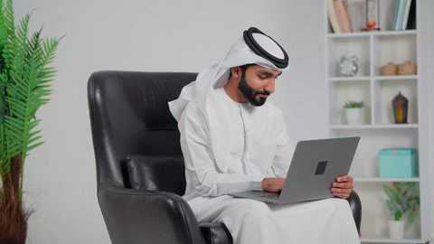 Communication and chatting with friends and family via social media, accomplishing and managing tasks online, the concept of remote work, integrating work with modern and advanced technology, an Arab Gulf Emirati man wearing a kandura and ghutrah sitting on a chair using a laptop.