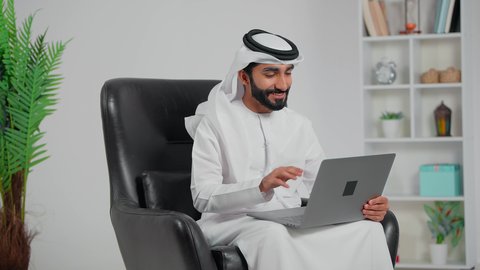 مفهوم القبول و الموافقة، إجراء مكالمة فيديو بإستخدام جهاز الحاسوب المحمول،  رجل عربي خليجي اماراتي يرتدي الكندورة و الغتره  يجلس على كرسي يقوم عقد إجتماع عمل عبر اللابتوب، رفع كف اليد بايماءات التحية و السلام