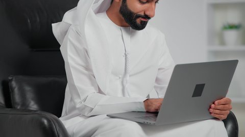 انجاز وادارة المهام عبر الأونلاين، مفهوم العمل عن بعد, دمج العمل مع التقنية والتكنولوجيا الحديثة والمتطورة,  تصوير مقرب لرجل عربي خليجي اماراتي يرتدي الكندورة و الغتره يجلس على كرسي يستخدم الحاسب المحمول, التواصل والدردشة مع الأصدقاء والأهل عبر السوشال ميديا