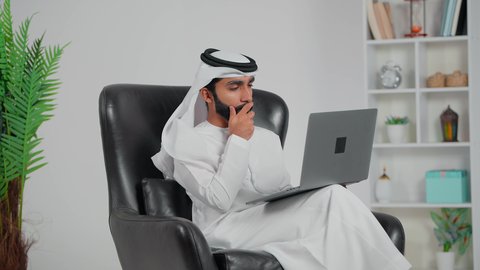 ايماءات الاندماج و التفكير فى شيء ما، مفهوم العمل عن بعد, دمج العمل مع التقنية والتكنولوجيا الحديثة والمتطورة, انجاز وادارة المهام عبر الأونلاين, رجل عربي خليجي اماراتي يرتدي الكندورة و الغتره يجلس على كرسي يستخدم الحاسب المحمول, التواصل والدردشة مع الأصدقاء والأهل عبر السوشال ميديا