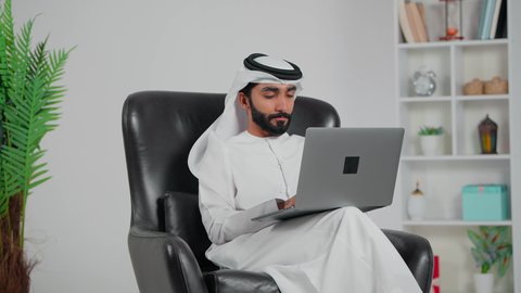 مفهوم العمل عن بعد, دمج العمل مع التقنية والتكنولوجيا الحديثة والمتطورة, انجاز وادارة المهام عبر الأونلاين, رجل عربي خليجي اماراتي يرتدي الكندورة و الغتره يجلس على كرسي يستخدم الحاسب المحمول, التواصل والدردشة مع الأصدقاء والأهل عبر السوشال ميديا, ايماءات الاندماج و التفكير فى شيء ما