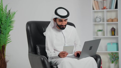 Integrating work with technology and tech, the skill of fast typing on the keyboard, an Arab Gulf Emirati man wearing a kandura and ghutrah sitting on a chair holding a notebook working on a laptop, performing administrative office tasks and functions, data entry and typing through the keyboard.