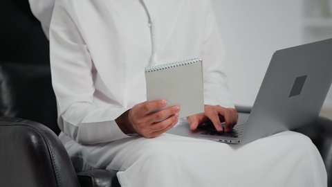 Integrating work with technology and tech, the skill of fast typing on the keyboard, an Arab Gulf Emirati man wearing a kandura and ghutrah sitting on a chair holding a notebook working on a laptop, performing administrative office tasks and functions, data entry and typing through the keyboard.