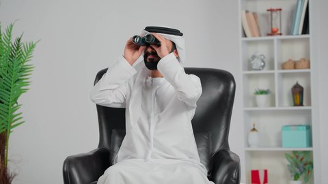 استخدام الأجهزة الحديثة لرؤية الأشياء البعيدة،  التخطيط المستقبلي للأهداف والطموحات، حب الاستكشاف والمغامرة، رجل عربي خليجي اماراتي يرتدي الكندورة و الغتره يجلس على كرسي يحمل بيده المنظار ثنائي العينين ينظر من خلاله إلى شيء ما