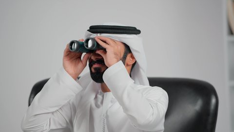 التخطيط المستقبلي للأهداف والطموحات، استخدام الأجهزة الحديثة لرؤية الأشياء البعيدة، حب الاستكشاف والمغامرة، تصوير مقرب لرجل عربي خليجي اماراتي يرتدي الكندورة و الغتره يجلس على كرسي يحمل بيده المنظار ثنائي العينين ينظر من خلاله إلى شيء ما