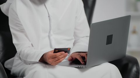 Payment through credit card, monitoring banking services through a laptop, purchasing through electronic applications, a close-up shot of an Emirati Gulf Arab man wearing a kandura and ghutrah sitting on a chair holding a bank card in his hands using a laptop.