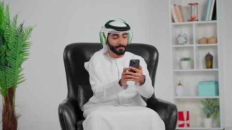 تصفح مواقع التواصل الاجتماعي، الاندماج والتفاعل مع الانغام الموسيقية، الإستماع للأغاني بإستخدام الهيدفون، رجل عربي خليجي اماراتي يرتدي الكندورة و الغتره يجلس على كرسي يضع سماعة رأس و يمسك الهاتف المحمول, مواهب غنائية فنية
