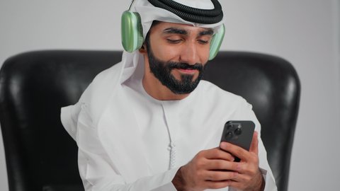 تعابير وايماءات السرور لسماع الأغنية، الاندماج والتفاعل مع الانغام الموسيقية، الإستماع للأغاني بإستخدام الهيدفون، تصوير مقرب لرجل عربي خليجي اماراتي يرتدي الكندورة و الغتره يجلس على كرسي يضع سماعة رأس و يمسك الهاتف المحمول , مواهب غنائية فنية