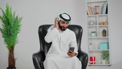 الاندماج والتفاعل مع الانغام الموسيقية، الإستماع للأغاني بإستخدام الهيدفون، تعابير وايماءات السرور لسماع الأغنية،رجل عربي خليجي اماراتي يرتدي الكندورة و الغتره يجلس على كرسي يضع سماعة رأس و يمسك الهاتف المحمول, مواهب غنائية فنية