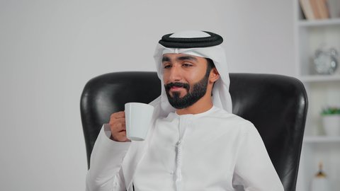 Sipping a hot drink, feeling refreshed and joyful, a close-up shot of an Emirati Gulf Arab man wearing a kandura and ghutrah sitting on a chair holding a cup of tea, enjoying moments of calm and comfort.