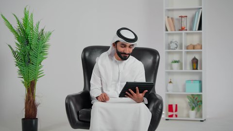 استخدام الاجهزة التقنية الحديثة، مفهوم إدارة المشاريع عن بعد،رجل عربي خليجي اماراتي يرتدي الكندورة و الغتره يجلس على كرسي  يقوم بانجاز مهام العمل عبر جهاز التابلت، تصفح مواقع و وسائل التواصل الاجتماعي