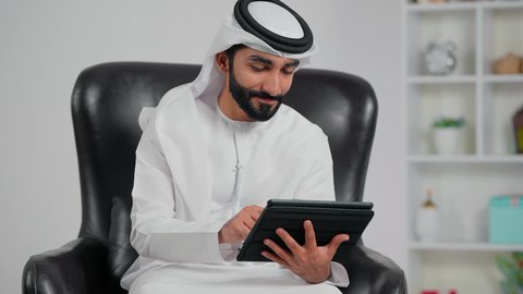 The concept of remote project management, an Arab Gulf Emirati man wearing a kandura and ghutrah sitting on a chair completing work tasks via a tablet, browsing websites and social media, using modern technological devices.
