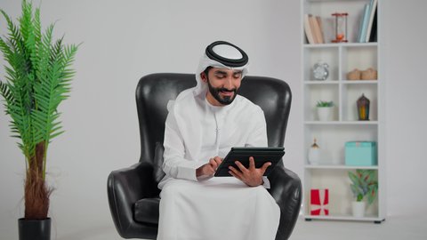 Gestures of happiness and joy, raising the hand with expressions of greeting and peace, an Arab Gulf Emirati man wearing a kandura and ghutrah sitting on a chair using a tablet to make a video call, accomplishing work tasks remotely.