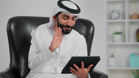 Raising the hand with expressions of greeting and peace, an Arab Gulf Emirati man wearing a kandura and ghutrah sits on a chair using a tablet to make a video call, accomplishing work tasks remotely, gestures of happiness and joy.