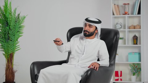 رجل عربي خليجي اماراتي يرتدي الكندورة و الغتره يجلس على كرسي يمسك جهاز التحكم عن بعد بالتلفاز , مشاهدة الأفلام والمسلسلات , البرامج الثقافية والأخبار , قضاء وقت الفراغ ,  مفهوم التسلية والترفيه