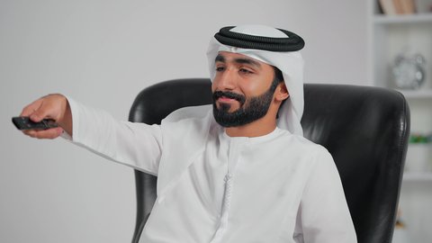 مشاهدة الأفلام والمسلسلات، رجل عربي خليجي اماراتي يرتدي الكندورة و الغتره يجلس على كرسي يمسك جهاز التحكم عن بعد بالتلفاز , البرامج الثقافية والأخبار , قضاء وقت الفراغ , مفهوم التسلية والترفيه