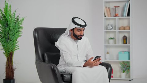 إجراء مكالمة فيديو،الدردشة مع الاهل و الاصدقاء ، جهاز تقني حديث ومتطور، رجل عربي خليجي اماراتي يرتدي الكندورة و الغتره يجلس على كرسي يقوم باستخدام الهاتف المحمول، تعابير الاندهاش و الذهول