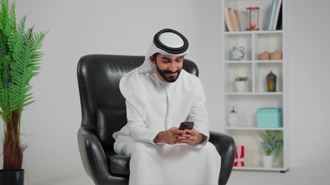 Chatting with family and friends, using a modern technological device, an Arab Gulf Emirati man wearing a kandura and ghutrah sitting on a chair holding a mobile phone, integrating technology and technology into daily life, browsing social media.