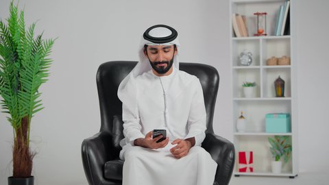 الاحتفاظ بالصور واللحظات التذكارية،رجل عربي خليجي اماراتي يرتدي الكندورة و الغتره يجلس على كرسي يقوم بالتقاط صورة سيلفي باستخدام الهاتف المحمول، استخدام التقنية الحديثة والمطورة، ايماءات السعادة والسرور