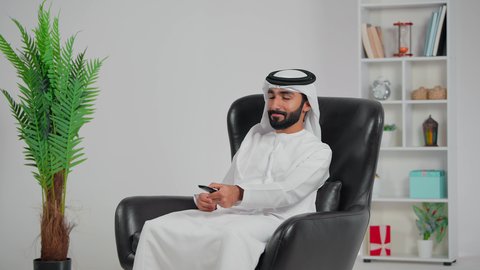 الشغف بمتابعه مباريات كرة القدم، رجل عربي خليجي اماراتي يرتدي الكندورة و الغتره يجلس على كرسي يقوم بمشاهده المبارة، رفع قبضة اليد بإيماءات الفوز والسرور،