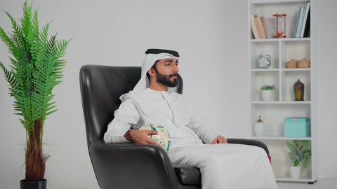 إيماءات الحزن لخسارة الفريق، متابعة مباريات كرة القدم، رجل عربي خليجي اماراتي يرتدي الكندورة و الغتره يجلس على كرسي يمسك بيدية الكرة ، تشجيع الأنديه والمنتخبات