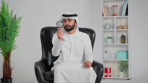 مفهوم الفحص والاستكشاف، توضيح الصورة والرؤية عن قرب، رجل عربي خليجي اماراتي يرتدي الكندورة و الغتره يجلس على كرسي يحمل عدسة مكبرة ينظر من خلالها بإيماءات التركيز والتمعن