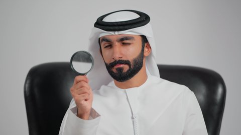 توضيح الصورة والرؤية عن قرب، مفهوم الفحص والاستكشاف،تصوير مقرب لرجل عربي خليجي اماراتي يرتدي الكندورة و الغتره يجلس على كرسي يحمل عدسة مكبرة ينظر من خلالها بإيماءات التركيز والتمعن