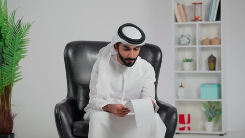 إيماءات التعجب والدهشة، التفاجؤ والاستغراب من شيء ما, خسارة وفقدان شيء معين، رجل عربي خليجي اماراتي يرتدي الكندورة و الغتره يجلس على كرسي يمسك ورق  و يضع يديه على رأسه بتعابير الصدمة والذهول