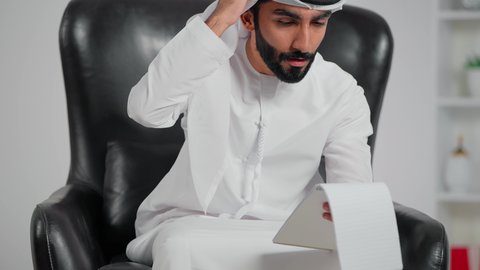 التفاجؤ والاستغراب من شيء ما, خسارة وفقدان شيء معين, إيماءات التعجب والدهشة،رجل عربي خليجي اماراتي يرتدي الكندورة و الغتره يجلس على كرسي يمسك ورق  و يضع يديه على رأسه بتعابير الصدمة والذهول