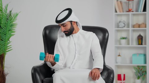 نشاط بدني للمحافظة على اللياقة الجسدية، رجل عربي خليجي اماراتي يرتدي الكندورة والغترة يجلس على كرسي و يمارس التمارين الرياضية مع الدمبل، كمال الأجسام و بناء العضلات، تمارين رفع الأثقال (دمبلز)، الأوزان الحديدية الثقيلة