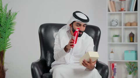 الاحتفال بأعياد الميلاد, التعبير عن المحبة والتقدير, تقديم الهدايا في المناسبات السعيدة, رجل عربي خليجي اماراتي يرتدي الكندورة و الغتره يجلس على كرسي يفتح صندوق هدايا بإيماءات المفاجاة والسرور