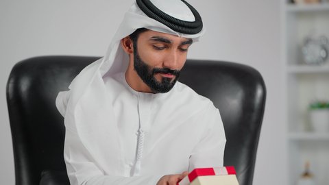التعبير عن المحبة والتقدير، الاحتفال بأعياد الميلاد, تقديم الهدايا في المناسبات السعيدة,  تصوير مقرب لرجل عربي خليجي اماراتي يرتدي الكندورة و الغتره يجلس على كرسي يفتح صندوق هدايا بإيماءات المفاجاة والسرور