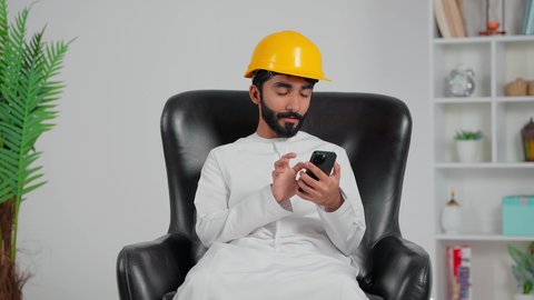 Chatting with family and friends, supervising and following up on projects, using modern technology, an Arab Gulf Saudi engineer wearing a white thobe and a safety helmet sitting on a chair using a mobile phone, integrating technology and tech into daily life, browsing social media.