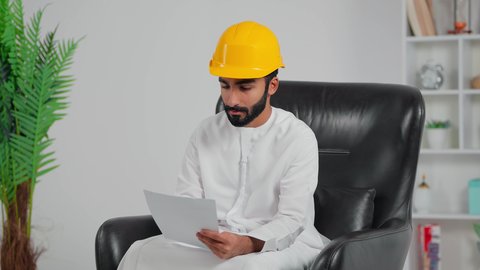 الإشراف على العمل وتدقيقه، إدارة ودراسة المشاريع الهندسية، متابعة سير المشروع في موقع العمل, مهندس عربي خليجي سعودي يرتدي الثوب الابيض و خوذة الحماية يجلس على كرسي و يقوم بقراء الملاحظات