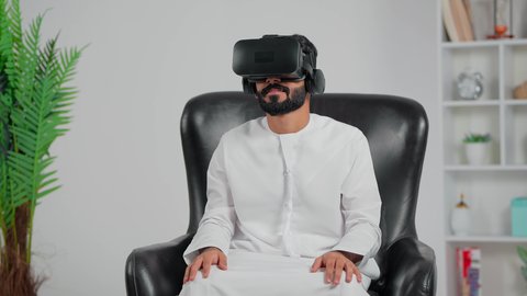 ايماءات الاندماج والتفاعل مع العالم الافتراضي، الاستمتاع في قضاء الأوقات الترفيهية عبر استخدام التقنيات الحديثة الالكترونية، ارجل عربي خليجي سعودي يرتدي الثوب الابيض يجلس على كرسي يضع نظارات الفي آر ثلاثية الابعاد، دمج التكنولوجيا بالحياة العملية