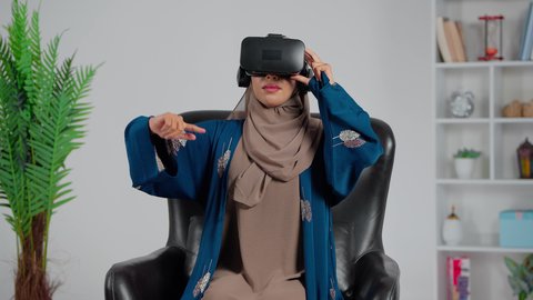 ايماءات الاندماج والتفاعل مع العالم الافتراضي، الاستمتاع في قضاء الأوقات الترفيهية عبر استخدام التقنيات الحديثة الالكترونية، امرأة عربية خليجية سعودية ترتدي العباءة والحجاب تجلس على كرسي تضع نظارات الفي آر ثلاثية الابعاد، دمج التكنولوجيا بالحياة العملية