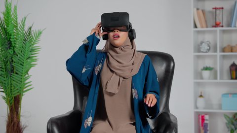 الاستمتاع في قضاء الأوقات الترفيهية عبر استخدام التقنيات الحديثة الالكترونية، امرأة عربية خليجية سعودية ترتدي العباءة والحجاب تجلس على كرسي تضع نظارات الفي آر ثلاثية الابعاد، ايماءات الاندماج والتفاعل مع العالم الافتراضي، دمج التكنولوجيا بالحياة العملية