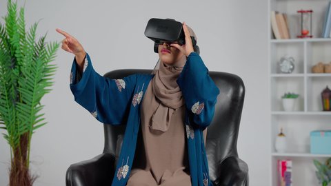 المتعة و التسلية بالمشاهدة عبر نظارة الواقع الافتراضي، تطور التقني والتكنولوجي، امرأة عربية خليجية سعودية ترتدي العباءة والحجاب تجلس على كرسي تنظر من خلال نظارات ثلاثية الأبعاد (الفي ار), الاندماج ومحاكاة العالم الخيالي،الاشارة الى شي ما