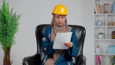 إدارة ودراسة المشاريع الهندسية، متابعة سير المشروع في موقع العمل, مهندسة عربية خليجية سعودية ترتدي العباءة و خوذة الحماية  تجلس على كرسي وتقوم بقراء الملاحظات، الإشراف على العمل وتدقيقه