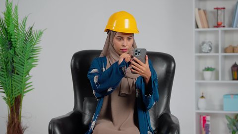 اشراف ومتابعة المشاريع، الدردشة مع الأهل والأصدقاء، استخدام جهاز تقني حديث، مهندسة عربية خليجية سعودية ترتدي العباءة والحجاب و خوذة الحماية تجلس على كرسي تمسك بيديها الهاتف المحمول، دمج التقنية والتكنولوجيا بالحياة اليومية، تصفح مواقع التواصل الاجتماعي
