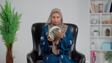 The concept of listening and hearing, a Gulf Arab Saudi woman wearing an abaya and hijab sitting on a chair with headphones on, listening to music, using headphone technology, enjoying her free time, gestures of happiness and engagement.